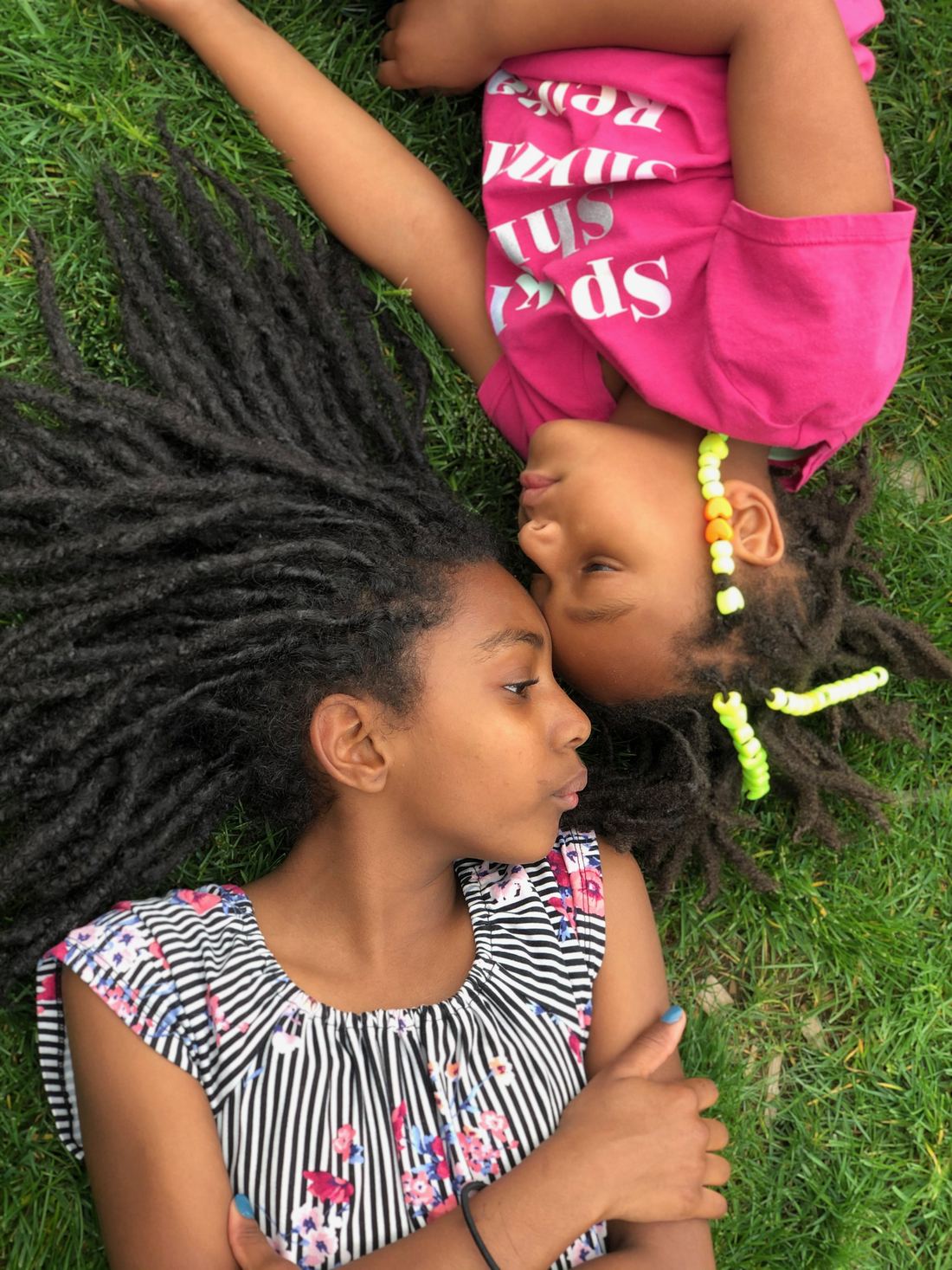 Two girls lying head to head on the grass, one with long locs fanned out, the other with beaded locs