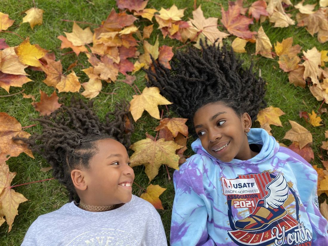 Two kids with locs lying in fallen autumn leaves, smiling at each other