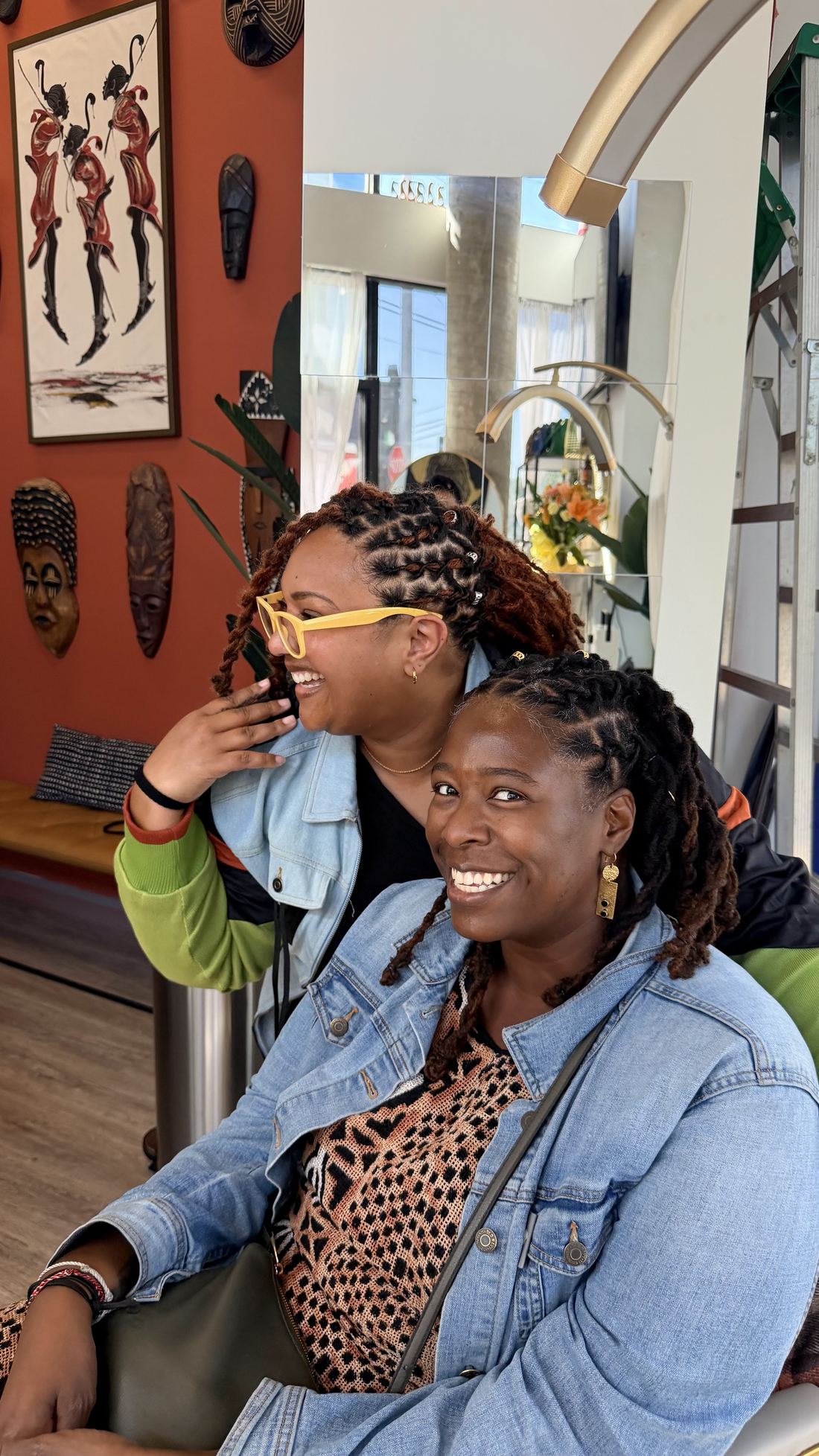 Two women laughing together in the studio, with African art on the terracotta wall behind them